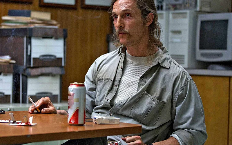 Matthew McConaughey as Rust sitting on a chair by a table with beer and cigarette
