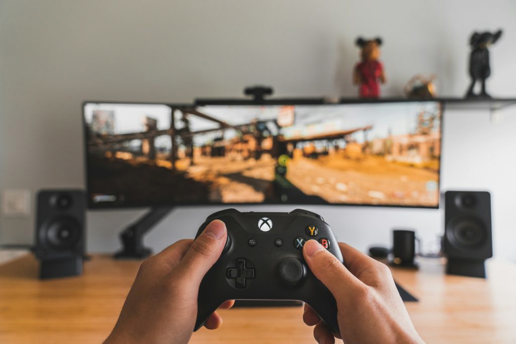A person holding up an Xbox controller while gaming in front of a screen.