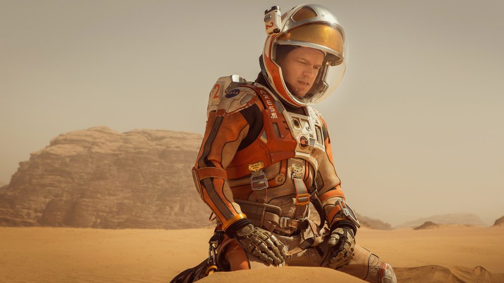 Ridley Scott directs The Martian. Matt Damon sits alone on the orange sand of Mars. He is in a space suit. 