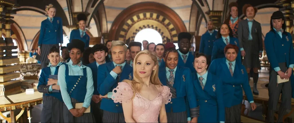 Ariana Grande as Glinda in Wicked, wearing a pink dress, surrounded by people wearing navy blue clothes. 