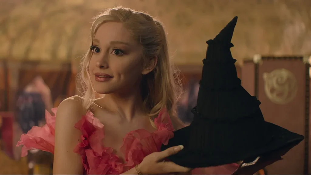 Ariana Grande holding up a black wizard hat as Glinda from Wicked. 