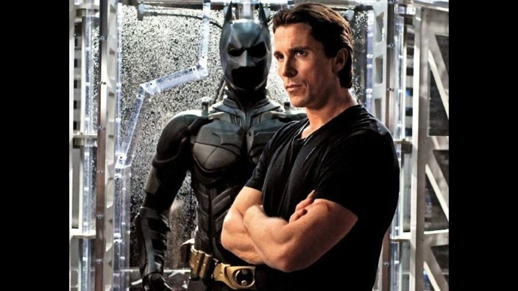 Christian Bale posing with his hands crossed in front of the Batman suit from Nolan trilogy. 