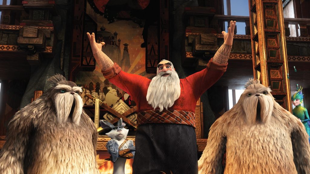 A scene inside North's workshop from 'Rise of the Guardians' showing North (Santa Claus) standing with his arms raised triumphantly. He's flanked by two large, furry Yeti workers, and a disgruntled-looking Easter Bunny.