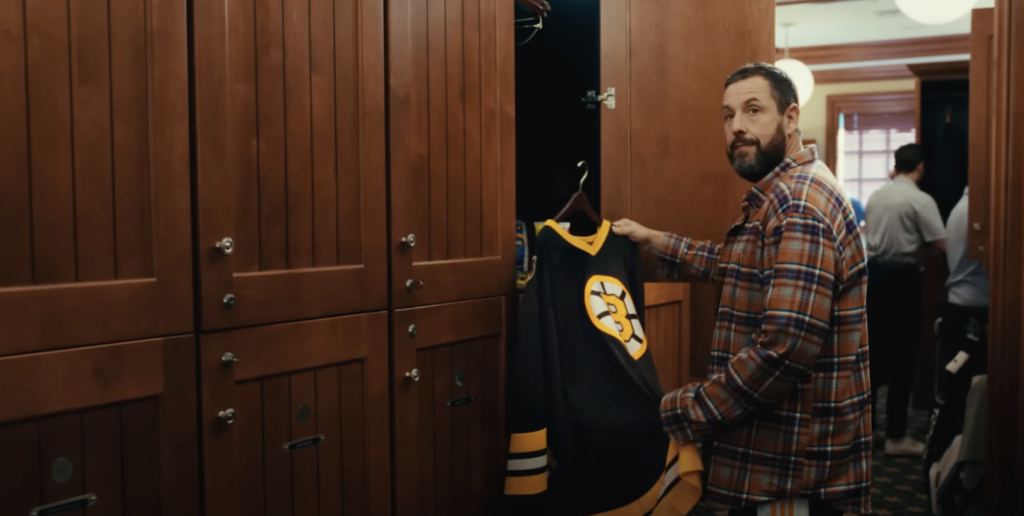 Adam Sandler in chequed shirt, holding a sport jersey. 