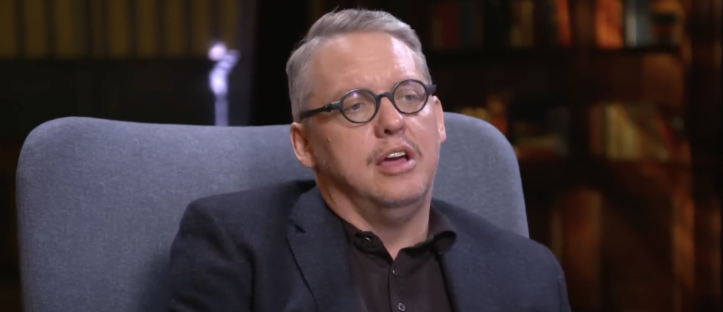 Adam McKay wearing a black suit in an interview. 