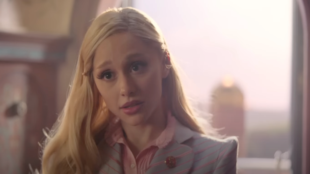 Ariana Grande with blonde hair, gray suit and pink shirt in Wicked, as Glinda. 