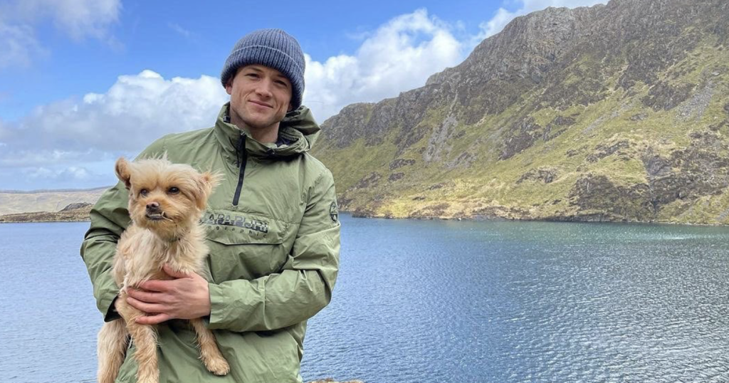 Taron Egerton holding a dog and posing for a picture at a scenic location. 