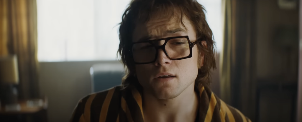 Taron Egerton as Elton John in Rocketman, wearing square framed glasses and stripped shirt. 