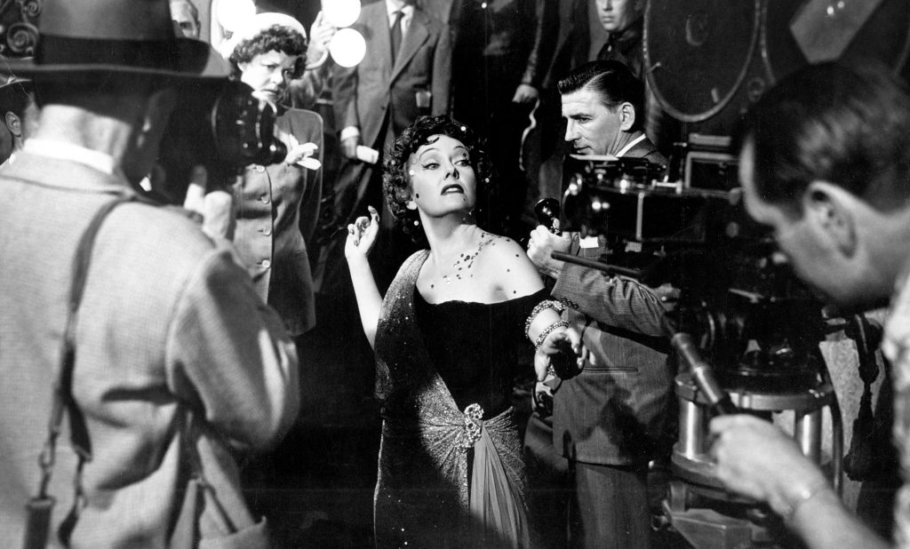 A black and white picture of Gloria Swanson as Norma Desmond in a still from the 1950 movie "Sunset Boulevard." She's striking a theatrical pose while being surrounded by film crew members, including cameramen with vintage equipment and other production staff and celebrities in suits.