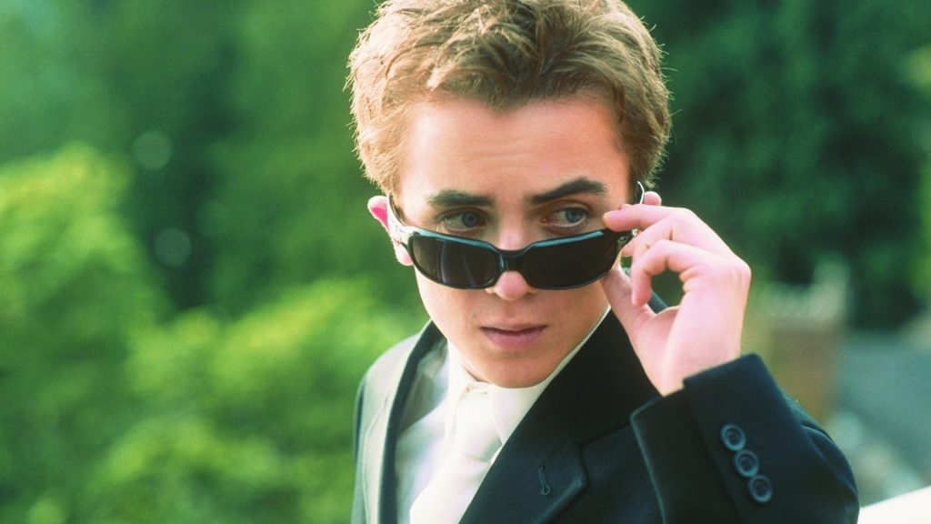 A close-up shot of Frankie Muniz as Cody Banks in a still from the 2003 movie "Agent Cody Banks." He's peering over dark sunglasses with his hand on the frame.