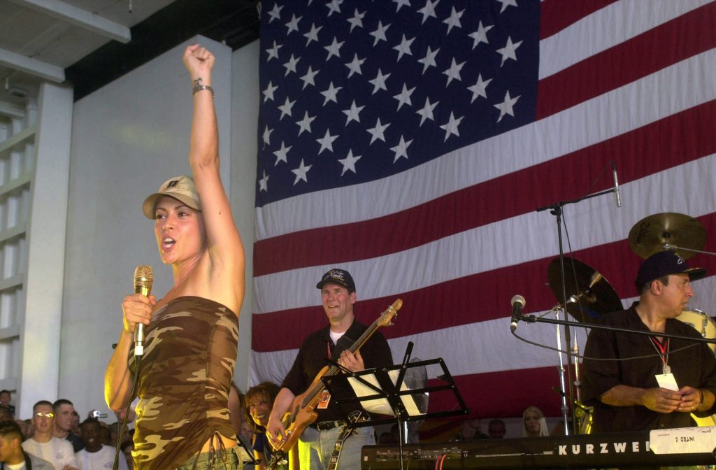 Actress Alyssa Milano speaks to crewmembers during a visit aboard USS Nimitz (CVN 68) for a United Services Organization (USO) show. She is in a camouflage attire and has raised her fist energetically.