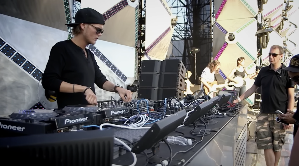 Avicii wearing a black shirt and cap, standing behind his DJ booth. 