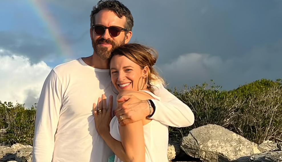Ryan Renolds embracing Blake Lively while both are smiling at the camera. There is a rainbow visible in the background.