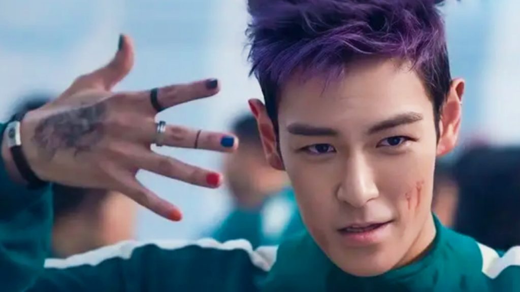 T.O.P with purple hair in Squid Game, holding up five fingers 