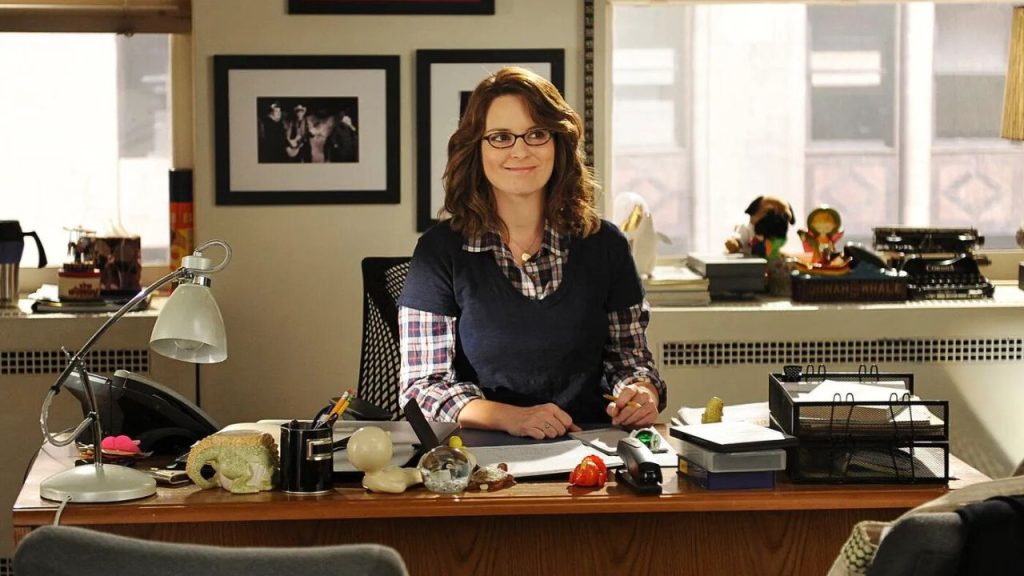 Tina Fey sitting at her office desk and smiling, in 30 Rock 