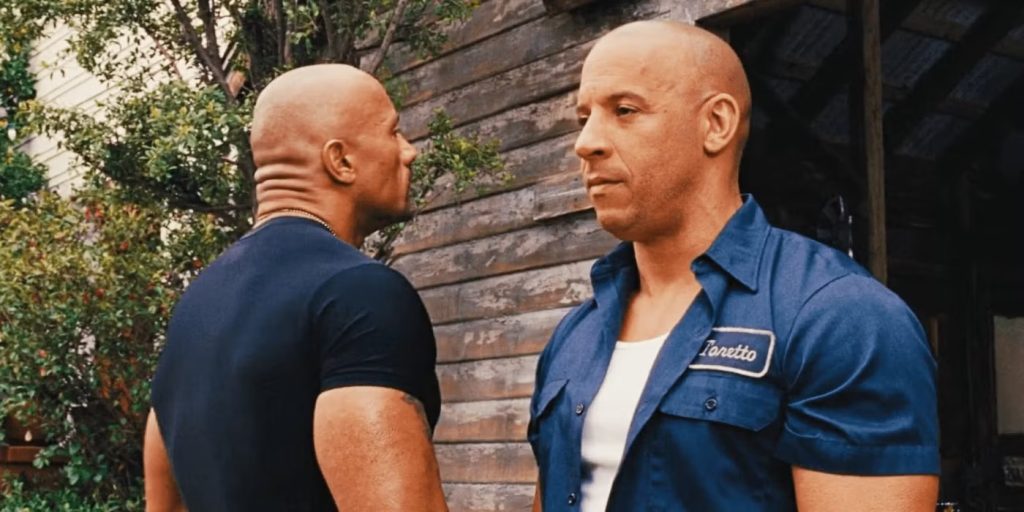 A scene from Fast & Furious 6 showing Vin Diesel as Dominic "Dom" Toretto and Dwayne "The Rock" Johnson as Luke Hobbs having an intense conversation against a wooden wall backdrop with foliage visible.