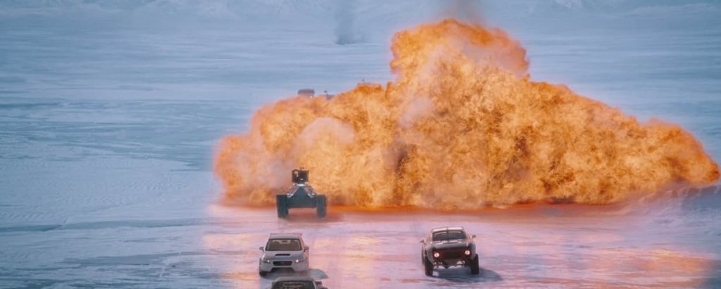 A dramatic action scene from The Fate of the Furious showing multiple vehicles racing across what appears to be an icy or snowy terrain. A massive orange explosion erupts behind them, with one vehicle being launched into the air by the blast. The scene is captured in a cinematic wide shot.