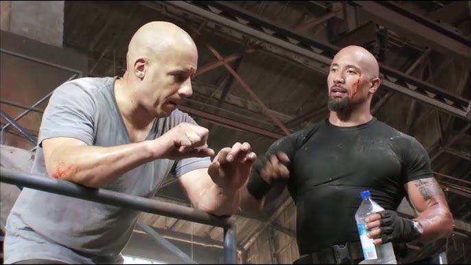 An industrial setting from Fast & Furious 6 showing Vin Diesel as Dominic "Dom" Toretto and Dwayne "The Rock" Johnson as Luke Hobbs engaged in conversation.