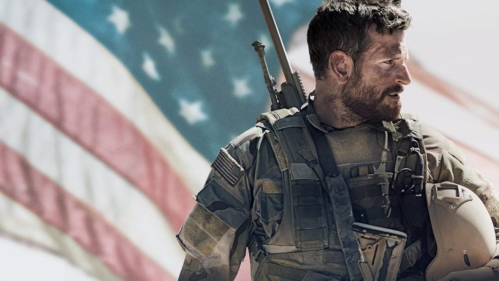 Bradley Cooper walking with a sniper on his back and a helmet in his hand while the flag of America waves behind him in the air.