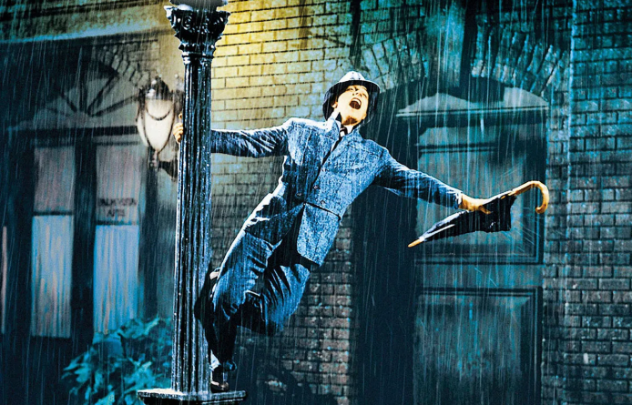 Gene Kelly dancing in the rain as Don Lockwood in the musical romantic comedy movie Singin’ in the Rain wearing a suit and carrying an umbrella.