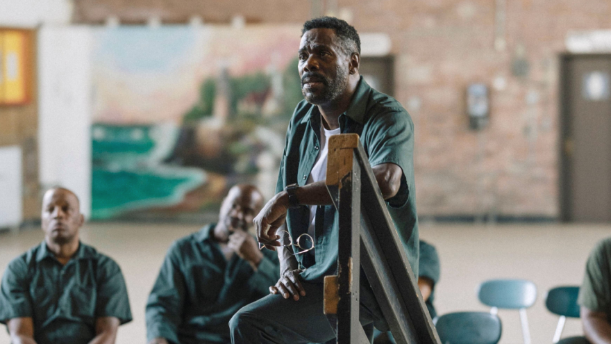 A scene from "Sing Sing" where Colman Domingo as Divine G in a dark green prison uniform stands in a communal space within a correctional facility. He is leaning against a piece of furniture. In the background, other individuals in similar green uniforms are sitting and watching.