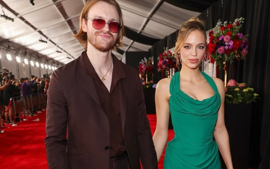 Claudia Sulewski wearing a brown suit with partner Finneas in a green dress at Grammy 2025.