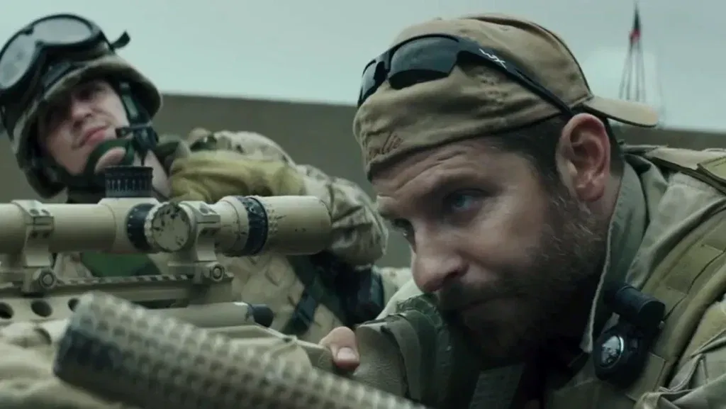 Bradley Cooper's Chris Kyle looking through his sniper scope while prone while a shooter smiles to him in the backdrop in Clint Eastwood's American Sniper.