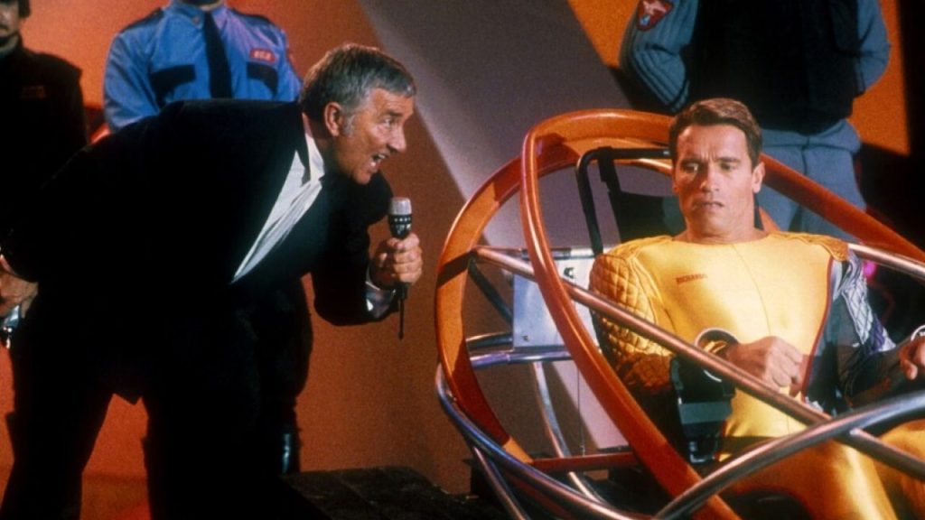 Richard Dawson speaking into a microphone and Arnold Schwarzenegger looking worried in The Running Man (1987)