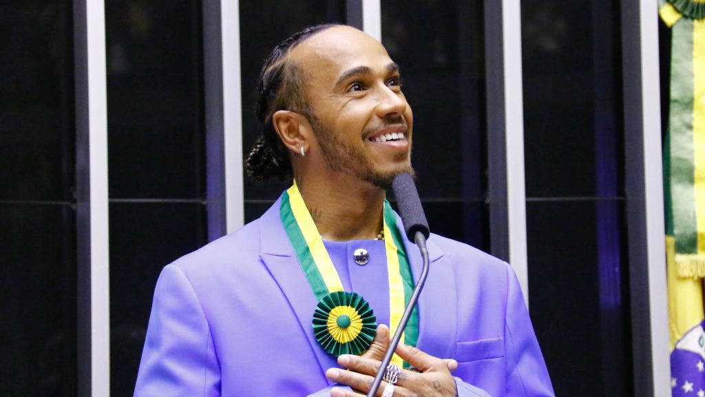 Lewis Hamilton smiling while wearing a purple suit jacket and a green and gold medallion. He is standing at a podium with a microphone, appearing to be at an awards ceremony.