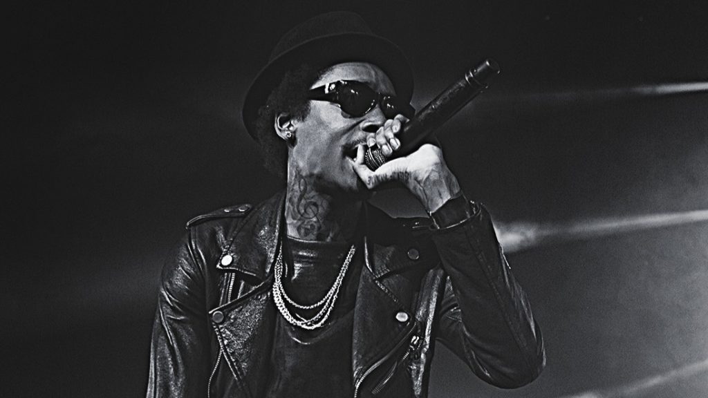 A black and white performance photo of Wiz Khalifa on stage. He is wearing a leather jacket, hat, and chains while singing into a microphone.