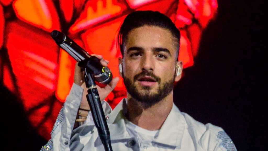 A performance shot of Maluma on stage wearing a white outfit with embellished details. He is holding a microphone and performing against a red-lit background.