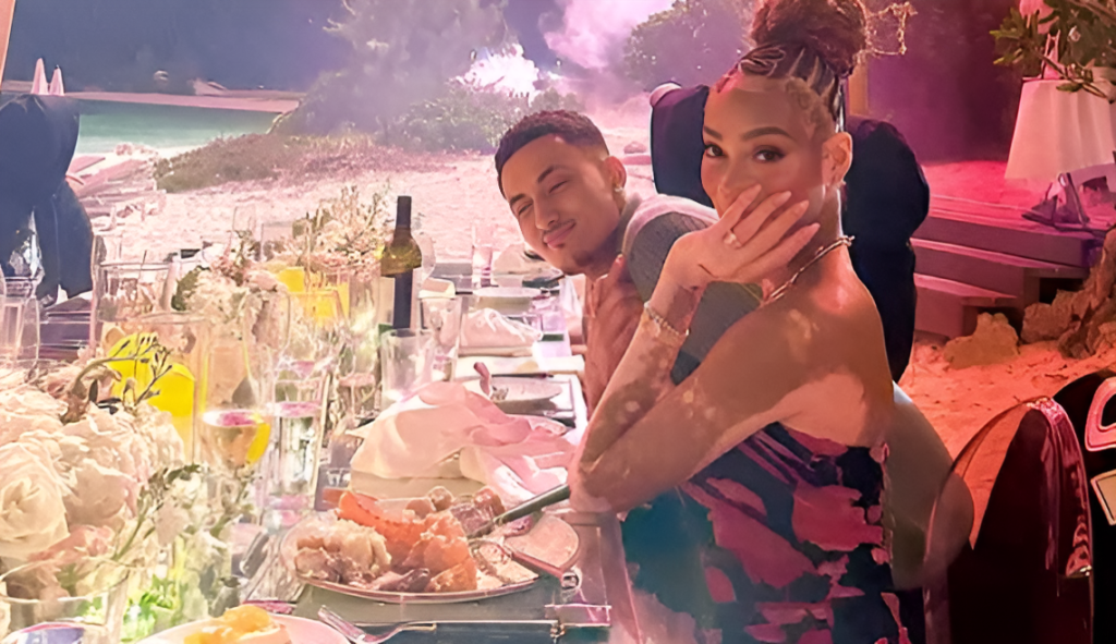 A candid moment showing Kyle Kuzma and Winnie Harlow at what appears to be a dinner setting. Harlow is covering her mouth while smiling, wearing an elegant dark dress, while Kuzma is seen smiling in a gray top. A decorated table with food and drinks is visible in the foreground.