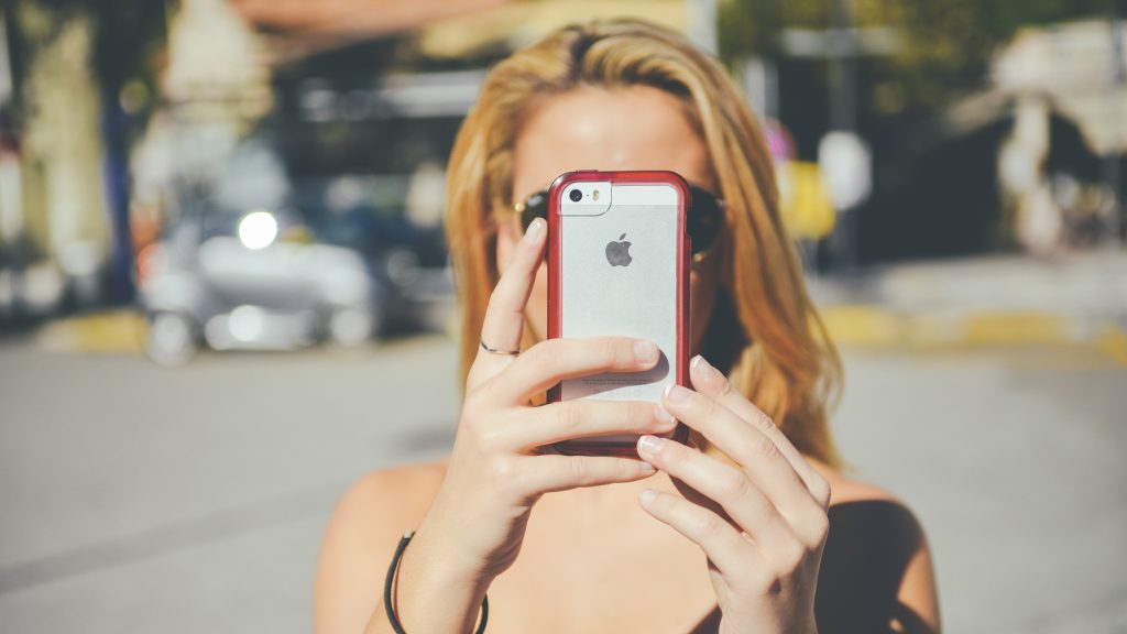 A person with blonde hair holds up an iPhone 5/5S with a red case, photographing something in front of them. The phone's distinctive Apple logo is visible through the clear portion of the case.