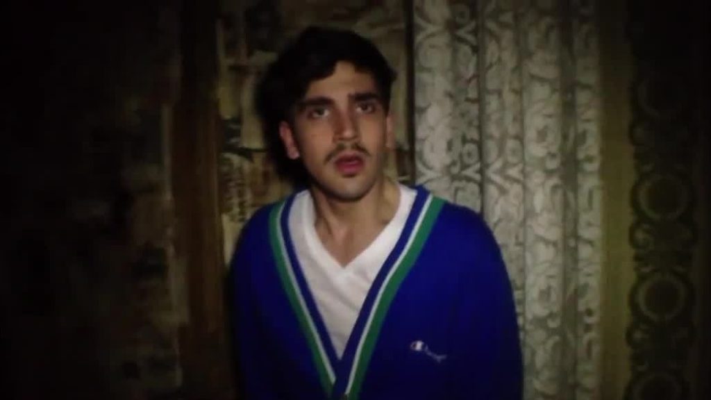 A close-up shot of Jonah Ehrenreich in a blue and white striped sweater against a patterned curtain background in low lighting.