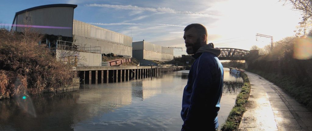 Anthony Mark Streeter in a blue hoodie standing by a canal or waterway at sunset, with industrial buildings and a bridge visible in the background.