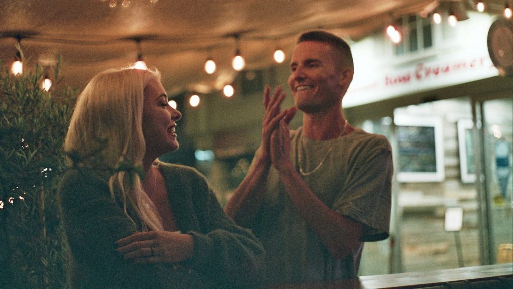 A nighttime scene of Bella Claire Martinez and Cal Barnes laughing together under string lights. Both appear to be sharing a joyful moment.