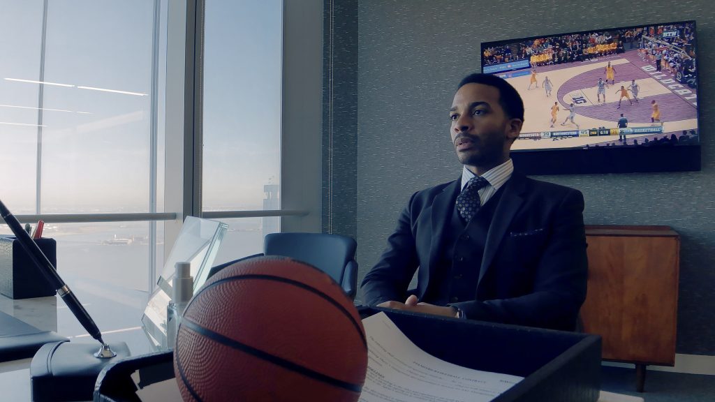 'André Holland' in a suit sitting at a desk with a basketball in the foreground and a basketball game playing on a TV screen in the background.