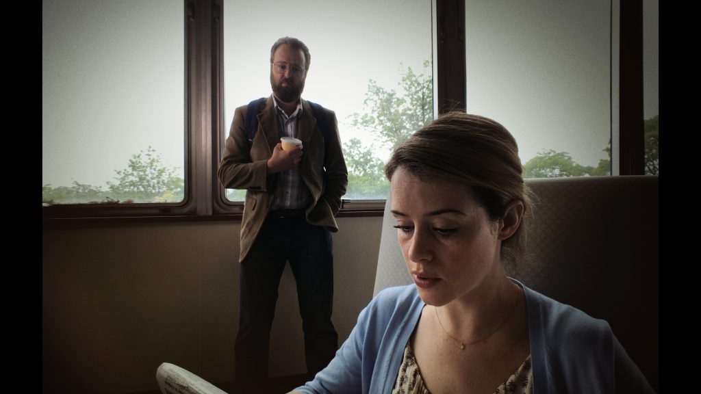 A cinematic shot showing Joshua Leonard in a brown coat standing while Claire Foy in light clothing sits, looking downward.