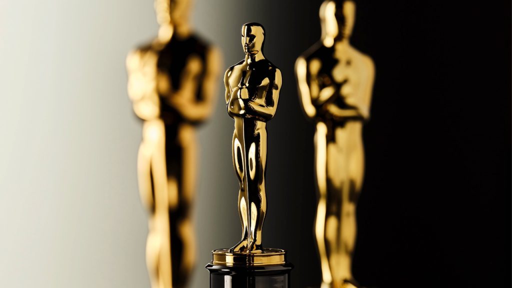 A close-up photograph of Oscar statuettes arranged in a row with selective focus. The center Oscar statue is in sharp focus while others are slightly blurred in the foreground and background.