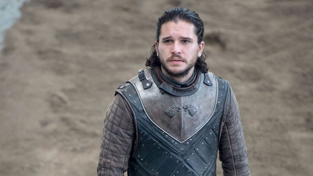 Jon Snow in Stark armor bearing the direwolf sigil. He's wearing battle gear with his hair pulled back, standing in what appears to be a battlefield or training ground. He has a solemn expression.