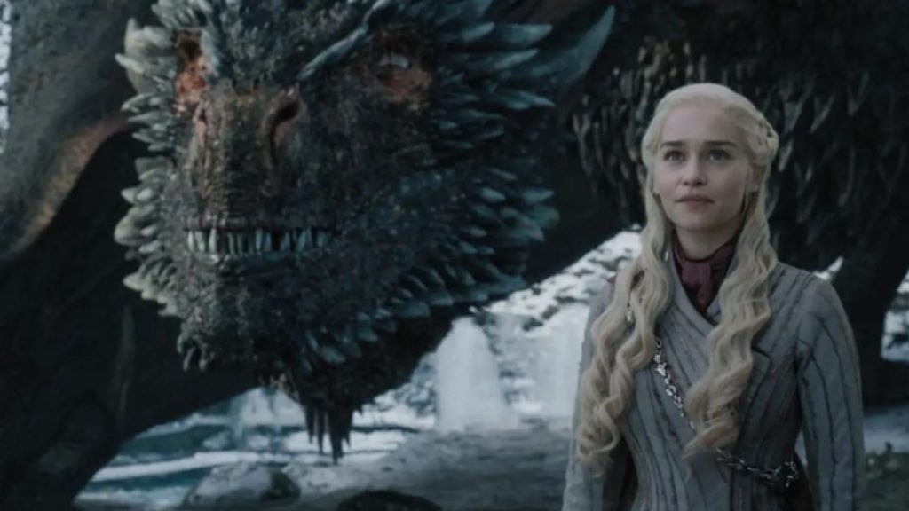 Daenerys Targaryen standing beside one of her dragons. She's wearing a light-colored winter outfit with her platinum blonde hair in her signature style. The dragon's detailed scales and face are visible in the foreground.