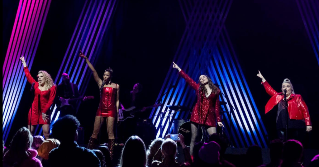 Busy Philipps, Renée Elise Goldsberry, Paula Pell, and Sara Bareilles performing on stage in red and black dresses, singing together in a scene.