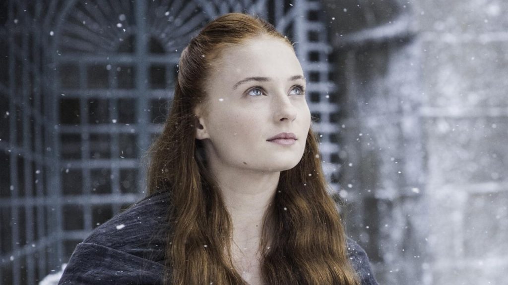 Sansa Stark in a winter scene with snowflakes visible around her. She has her distinctive auburn hair and is wearing a dark outfit, looking upward with a contemplative expression.