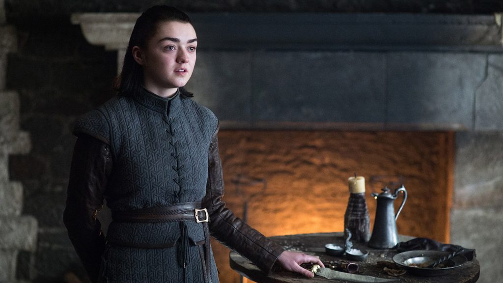 Arya Stark in her later seasons appearance, wearing Stark-styled battle dress with a leather tunic and belt. She's standing near a table with serving implements. The setting appears to be within a Northern castle interior.