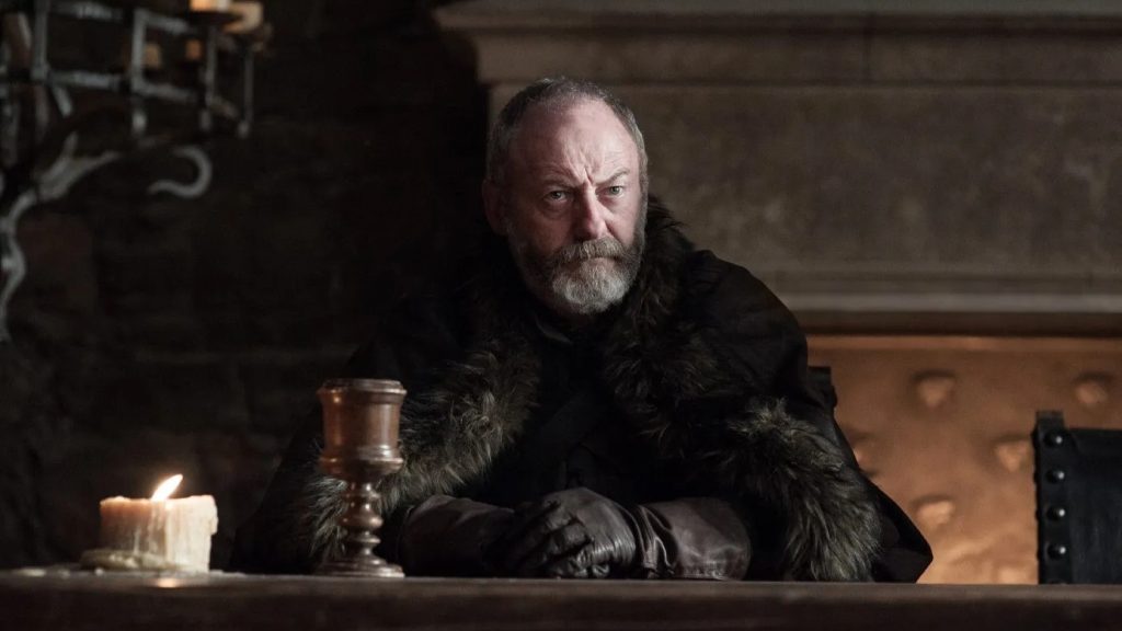 Davos Seaworth (the Onion Knight) seated at what appears to be a council table with a goblet nearby. He's wearing his typical Northern-style clothing with fur collars.