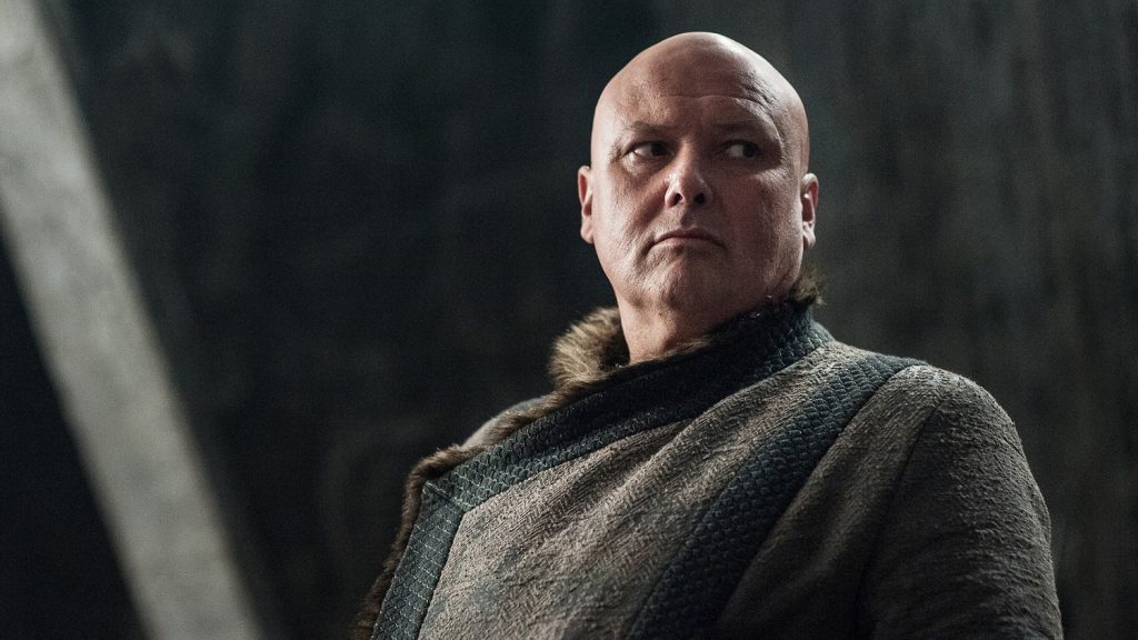 Varys (the Spider) in a dimly lit setting, showing his bald head and thoughtful, calculating expression. He's wearing the dark robes typical of his character.