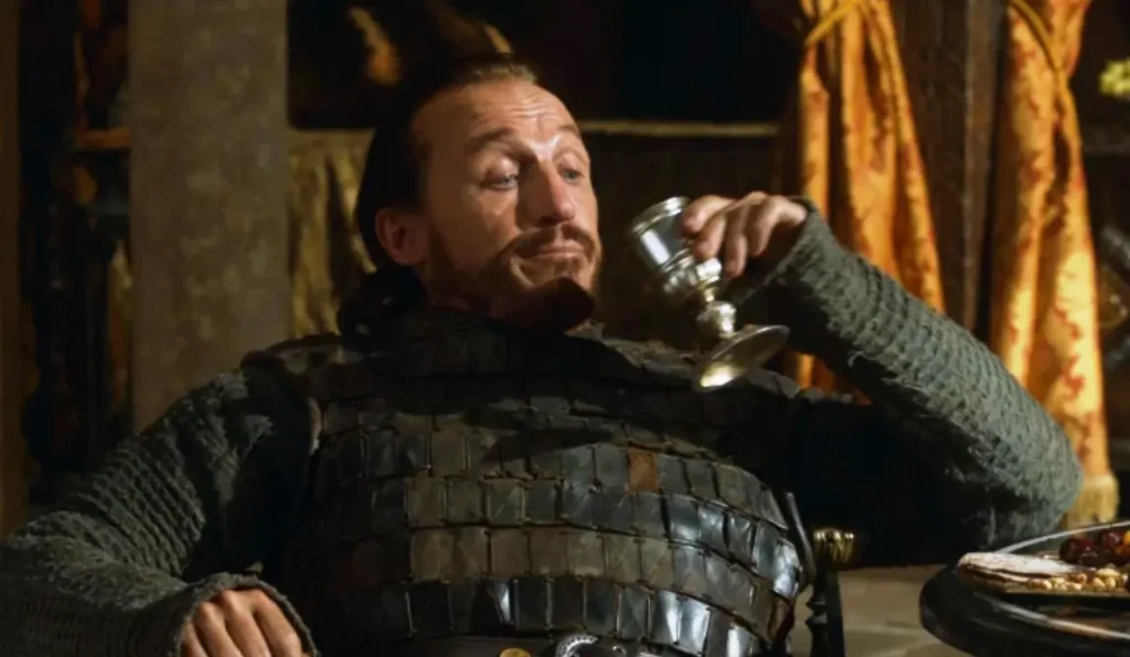 Bronn, the sellsword, enjoying a drink from a goblet while seated. He's wearing his characteristic armor with a smug, satisfied expression on his face.