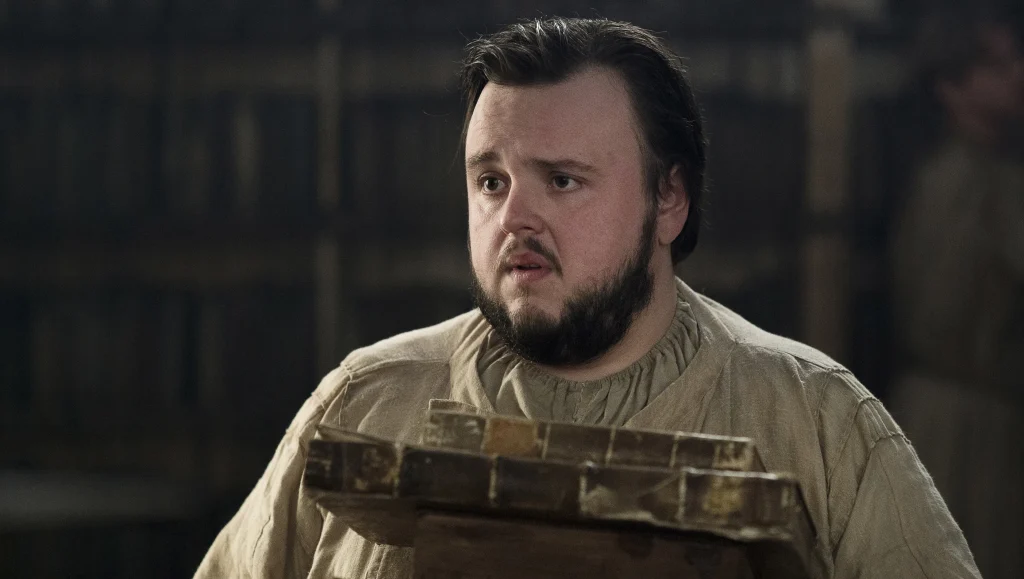 Samwell Tarly looking concerned while holding books. He's wearing simple clothing typical of his character at the Citadel.