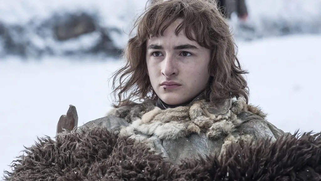 A close-up of Bran Stark in his Northern clothing, including heavy fur wrappings to protect from the cold. The image is set in a snowy landscape, showcasing his serious, distant expression.