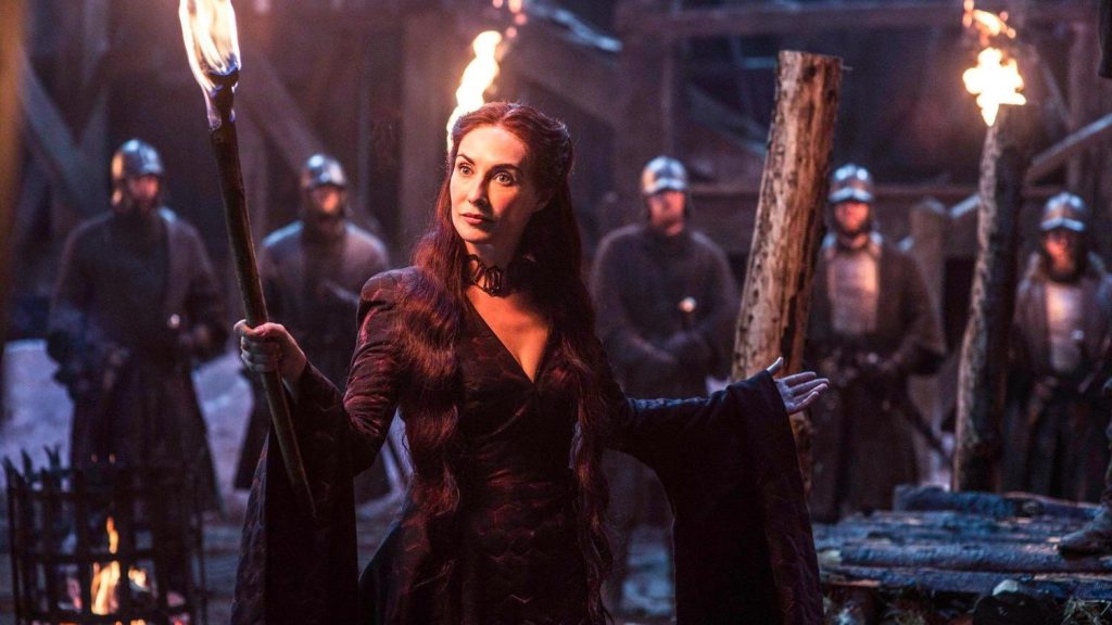 A scene from Game of Thrones showing Melisandre (the Red Woman) in a dark burgundy dress, holding a flaming torch while standing in a dimly lit castle setting. She has long red hair and a distinctive necklace, with armored guards visible in the background.